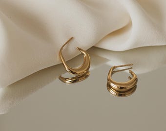 May include: A pair of gold hoop earrings with a curved, rounded shape. The earrings are on a white surface.