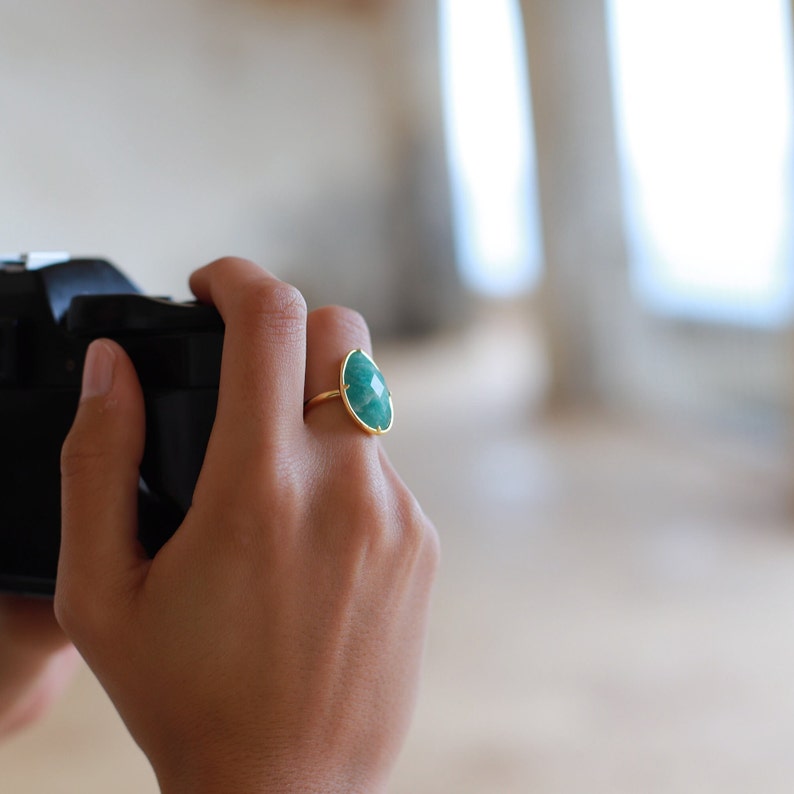 May include: A gold ring featuring a large, oval-shaped green gemstone. The ring is worn on a person's finger.
