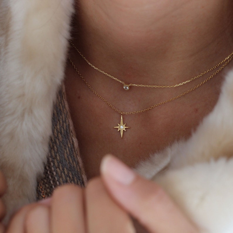 Puede incluir: Dos collares de oro con un colgante de estrella y un peque&ntilde;o colgante redondo. Los collares est&aacute;n superpuestos en el cuello de una persona.