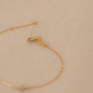 Op de afbeelding: Een delicate goudkleurige armband met een kleine kruisvormige hanger. De armband heeft een fijne ketting en een karabijnsluiting. De merknaam "ARAN" is zichtbaar op een klein label bij de sluiting. De achtergrond is een zachte, neutrale kleur.