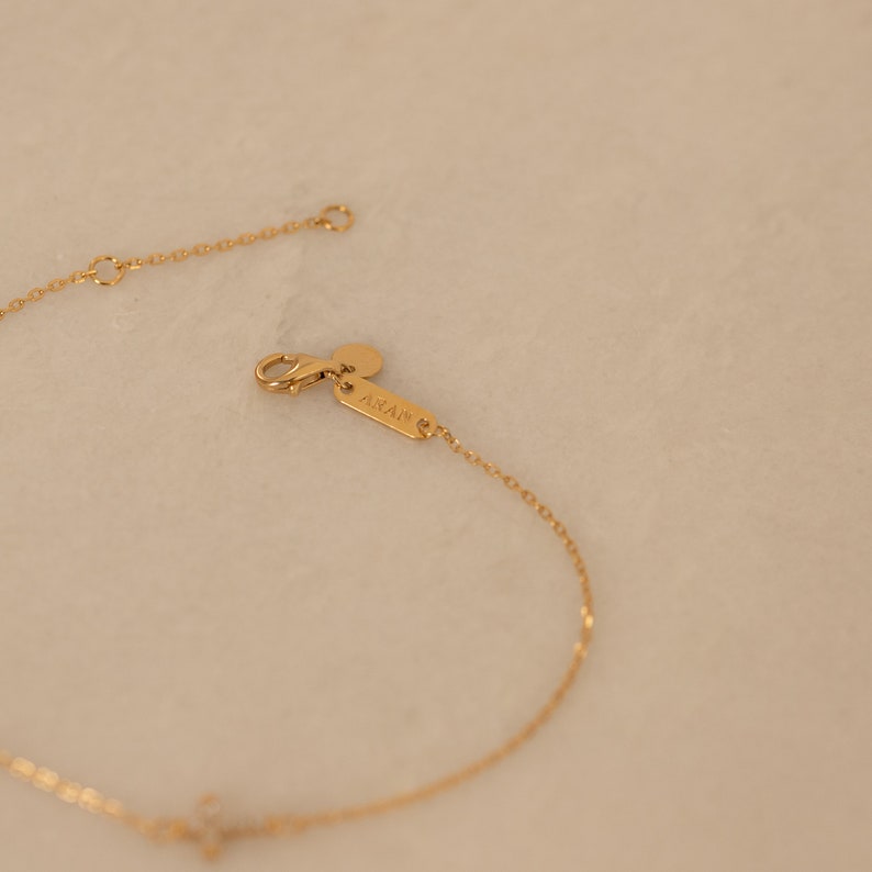Op de afbeelding: Een delicate goudkleurige armband met een kleine kruisvormige hanger. De armband heeft een fijne ketting en een karabijnsluiting. De merknaam "ARAN" is zichtbaar op een klein label bij de sluiting. De achtergrond is een zachte, neutrale kleur.