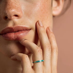 Può includere: Un anello in oro con intarsio turchese. L'anello è indossato su un dito, con una vista ravvicinata della mano e del viso. La pelle ha lentiggini e un incarnato naturale. Il design dell'anello è semplice ed elegante.