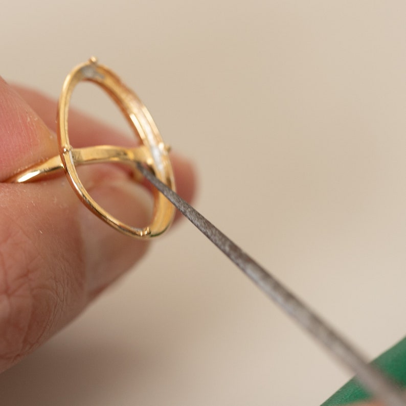 May include: A gold ring setting being filed with a metal file. The ring is being held by a hand and the file is being held by another hand.