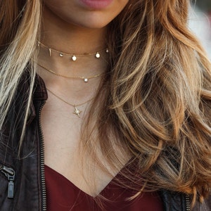 Puede incluir: Una mujer lleva una chaqueta de cuero marr&oacute;n y una blusa burdeos con tres collares de oro. Los collares tienen un colgante de estrella, un peque&ntilde;o diamante y dos peque&ntilde;os c&iacute;rculos.