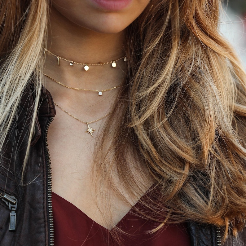 Puede incluir: Una mujer lleva una chaqueta de cuero marr&oacute;n y una blusa burdeos con tres collares de oro. Los collares tienen un colgante de estrella, un peque&ntilde;o diamante y dos peque&ntilde;os c&iacute;rculos.