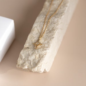 Puede incluir: Un collar de oro con un peque&ntilde;o colgante en forma de estrella incrustado con peque&ntilde;as piedras transparentes. El collar se muestra sobre una roca de color claro.