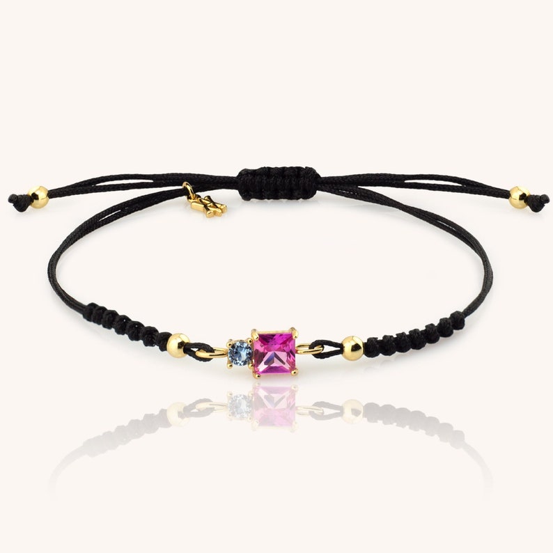 May include: A black string bracelet with a pink and blue gemstone charm. The charm is set in gold and features a square-shaped pink gemstone and a smaller round blue gemstone.