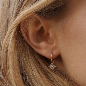 Puede incluir: Aretes de aro dorados con un pequeño dije de estrella colgante engastado con piedras de circonita cúbica transparentes.