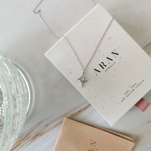 Puede incluir: Un collar de plata con un colgante en forma de estrella con una peque&ntilde;a piedra transparente. El collar est&aacute; en una caja blanca con el texto "ARAN JEWELS" y "925 SILVER JEWELS EST. 2014".