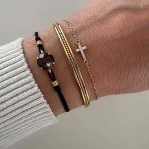 Op de afbeelding: Een verzameling gouden armbanden en een zwart koordarmband met een grote, facetgeslepen, bruine edelsteen. De gouden armbanden omvatten een armband en een delicate kettingarmband met een kruisvormige hanger.
