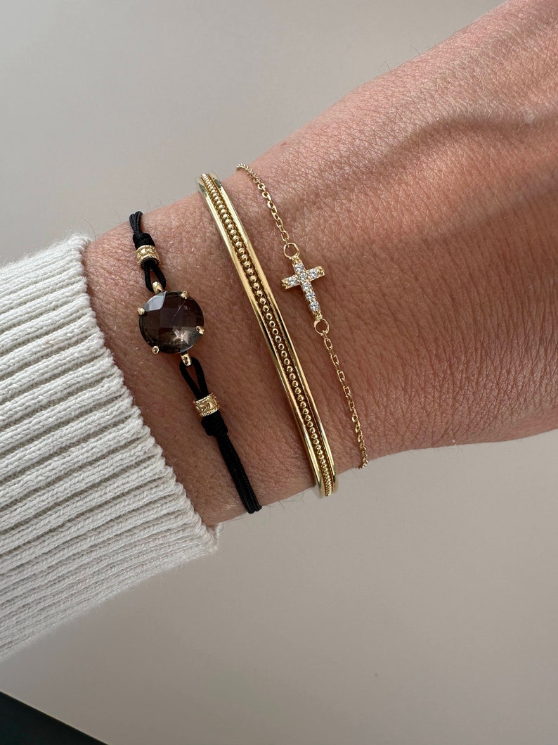 Op de afbeelding: Een verzameling gouden armbanden en een zwart koordarmband met een grote, facetgeslepen, bruine edelsteen. De gouden armbanden omvatten een armband en een delicate kettingarmband met een kruisvormige hanger.