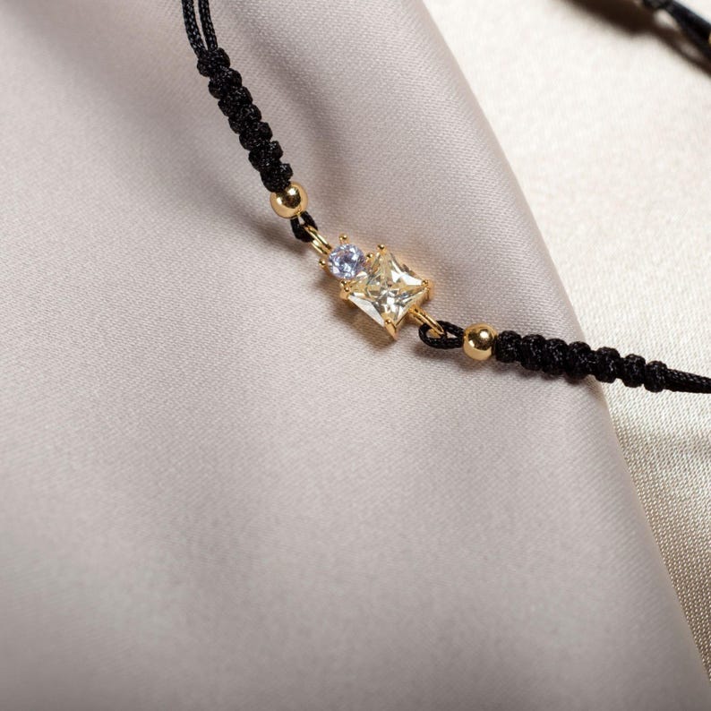 May include: A black cord bracelet with a gold square-shaped pendant featuring a clear crystal and a smaller purple crystal.