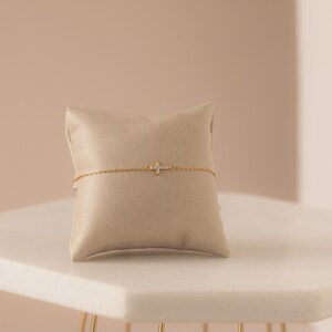 Op de afbeelding: Een delicate gouden armband met een kruisvormige hanger versierd met kleine, sprankelende edelstenen. De armband wordt gepresenteerd op een beige kussen, tegen een zachte, neutrale achtergrond. De kruishanger is het centrale punt en benadrukt het elegante ontwerp van de sieraden.