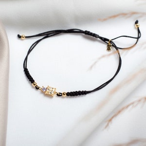 May include: A black string bracelet with a gold square-shaped charm featuring a clear crystal and a smaller purple crystal. The bracelet has a sliding knot closure.