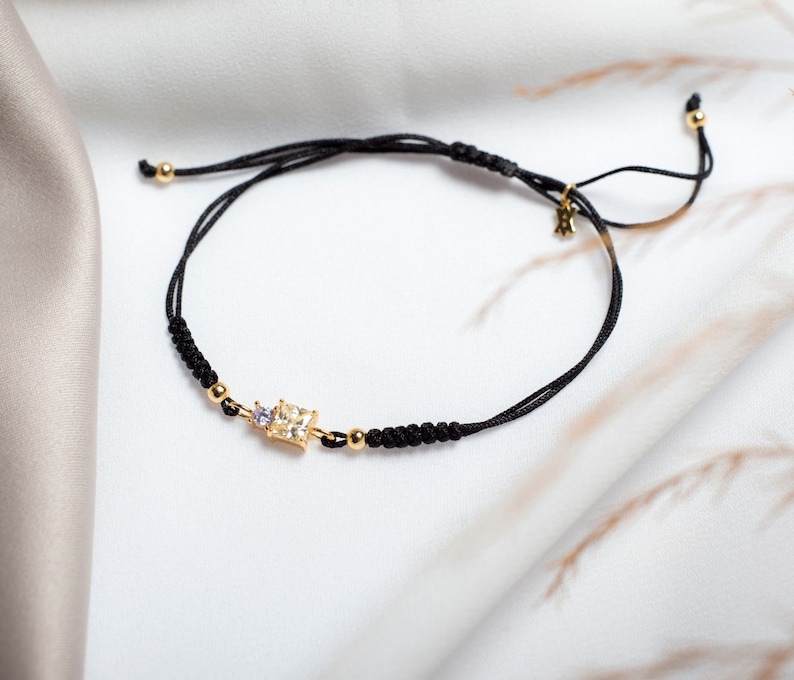 May include: A black string bracelet with a gold square-shaped charm featuring a clear crystal and a smaller purple crystal. The bracelet has a sliding knot closure.