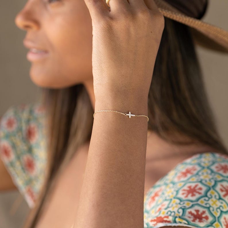 Op de afbeelding: Een fijne gouden armband met een kleine kruisbedel. De armband is slank en elegant, met het kruis in het midden van de pols. Dit sieraad is een eenvoudig maar stijlvol accessoire.
