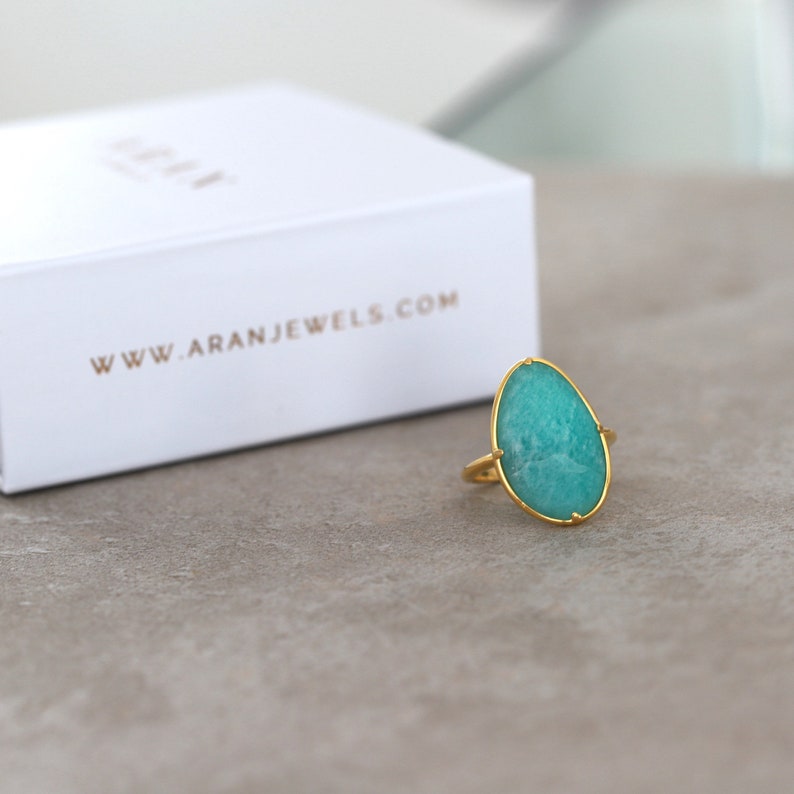 May include: A gold ring featuring a large, oval-shaped green gemstone. The ring is displayed on a grey surface.