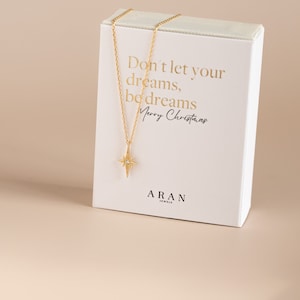 Puede incluir: Un collar de oro con un colgante de estrella y una peque&ntilde;a piedra transparente en el centro. El collar est&aacute; en una caja blanca con el texto "Don't let your dreams, be dreams Merry Christmas" y "ARAN JEWELS".