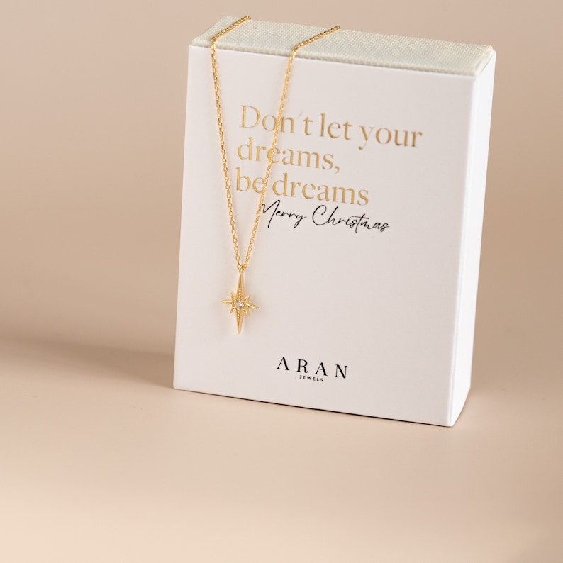 Puede incluir: Un collar de oro con un colgante de estrella y una peque&ntilde;a piedra transparente en el centro. El collar est&aacute; en una caja blanca con el texto "Don't let your dreams, be dreams Merry Christmas" y "ARAN JEWELS".