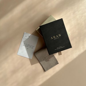 Op de afbeelding: Een bovenaanzicht van sieradenverpakkingen en een kleine kaart op een lichtbruin oppervlak. De zwarte doos heeft gouden letters met de tekst "ARAN JEWELS". Een kleine grijze pouch en een cr&egrave;mekleurige doos zijn ook zichtbaar. De kaart luidt "ALL YOU NEED IN LIFE IS A FRIEND WHO HAS CHOCOLATE... IT'S ME!"