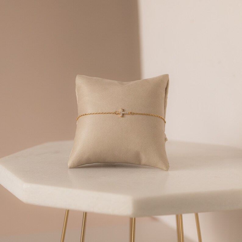 Op de afbeelding: Een goudkleurige armband met een kruisvormige hanger, versierd met kleine, heldere edelstenen. De armband wordt gepresenteerd op een beige kussen, tegen een neutrale achtergrond. De kruishanger is ongeveer 1,3 cm lang.