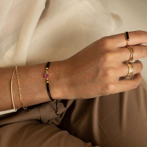 May include: A black string bracelet with a small pink gemstone and gold accents. The bracelet is tied with a knot and has a small gold bead at the end.