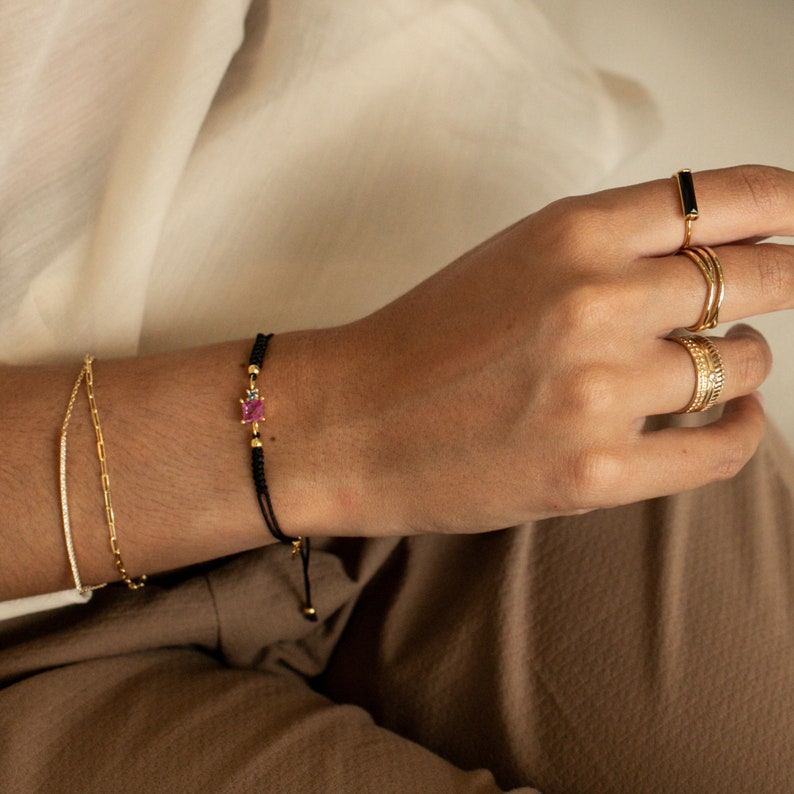 May include: A black string bracelet with a small pink gemstone and gold accents. The bracelet is tied with a knot and has a small gold bead at the end.