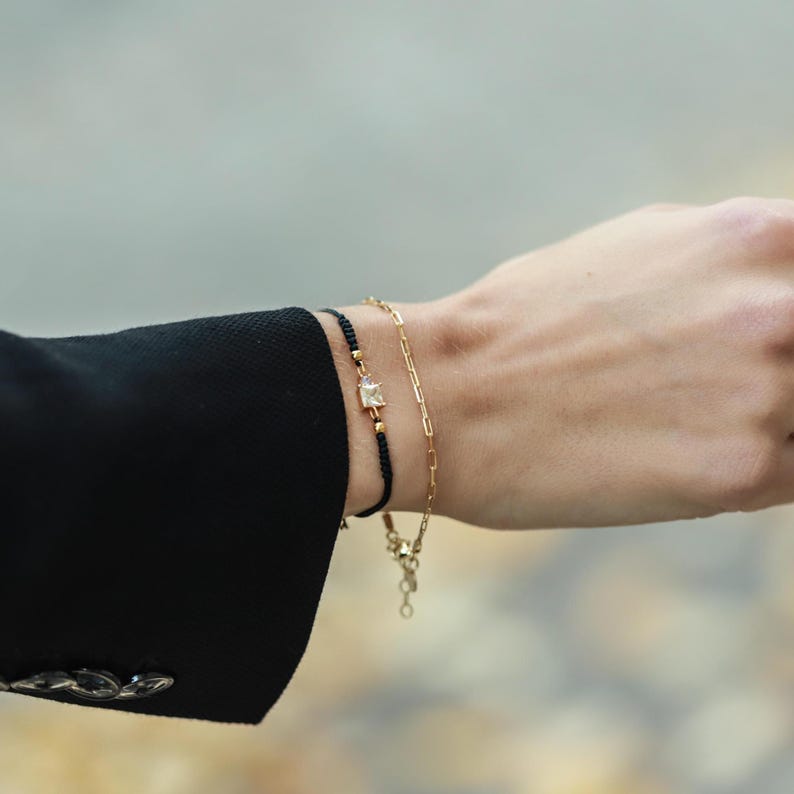 May include: Two bracelets on a wrist. One is a black string bracelet with a small gold charm with a clear stone. The other is a gold chain bracelet.