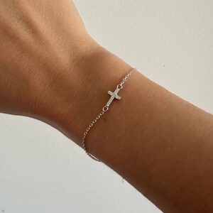 Op de afbeelding: Een delicate zilveren armband met een kleine kruisbedel versierd met sprankelende kristallen. De armband is bevestigd aan een fijne ketting, waardoor een minimalistisch en elegant ontwerp ontstaat. Het kruis is aan de zijkant van de pols geplaatst.