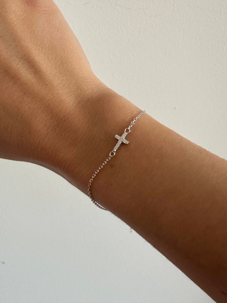 Op de afbeelding: Een delicate zilveren armband met een kleine kruisbedel versierd met sprankelende kristallen. De armband is bevestigd aan een fijne ketting, waardoor een minimalistisch en elegant ontwerp ontstaat. Het kruis is aan de zijkant van de pols geplaatst.