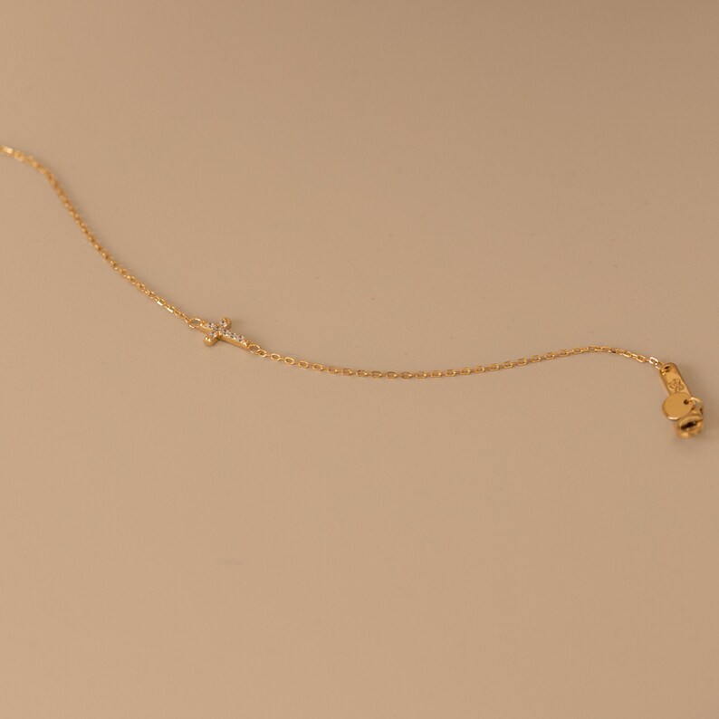 Op de afbeelding: Een delicate goudkleurige ketting met een kleine kruisvormige hanger versierd met sprankelende edelstenen. De ketting heeft een fijne ketting en een karabijnsluiting. De sieraden worden gepresenteerd tegen een neutrale achtergrond.