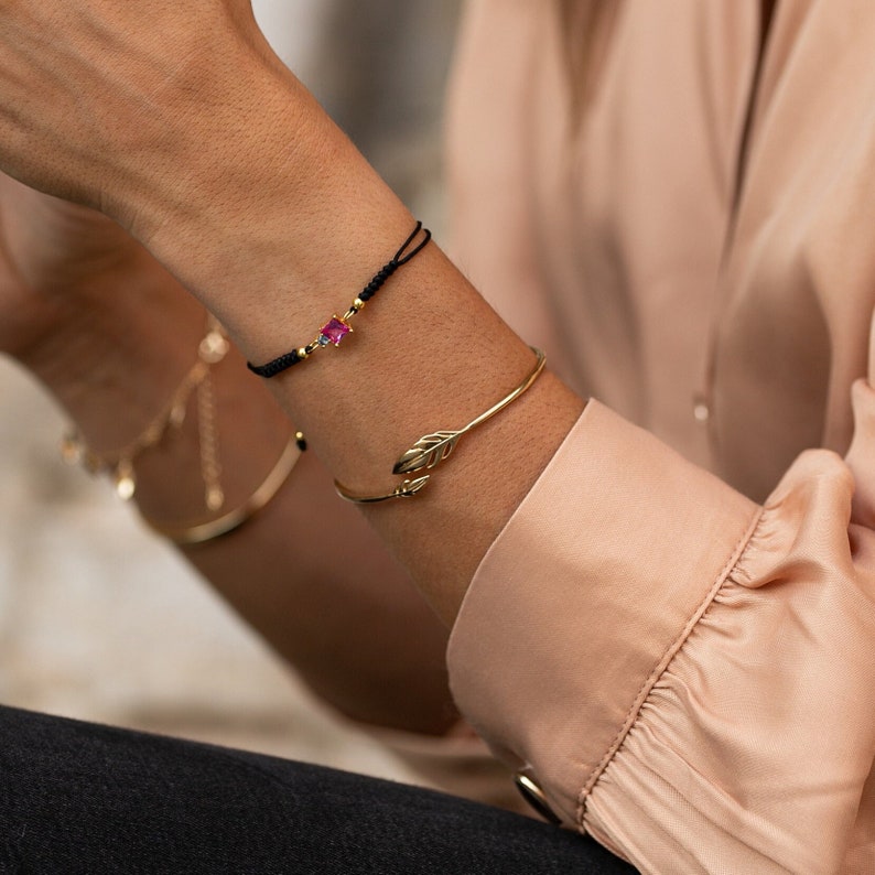 May include: A gold leaf bangle bracelet with a delicate design. The bracelet is worn on a wrist with a black string bracelet with a pink gemstone.