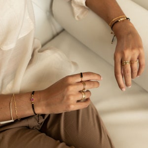 May include: A person wearing a variety of gold rings and bracelets. The bracelets are made of black string and gold beads. One bracelet has a pink stone charm. The rings are thin and delicate.
