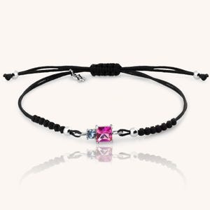 May include: A black string bracelet with a silver clasp and three gemstones. The gemstones are a pink square, a smaller blue round stone, and a smaller clear round stone.