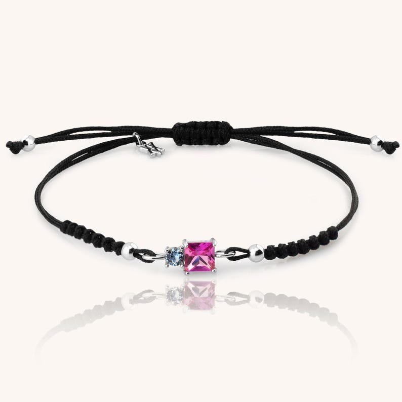 May include: A black string bracelet with a silver clasp and three gemstones. The gemstones are a pink square, a smaller blue round stone, and a smaller clear round stone.