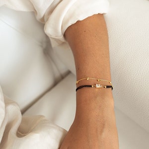 May include: Two bracelets on a wrist. The top bracelet is a delicate gold chain with small square-shaped stones. The bottom bracelet is a black string with a larger square-shaped stone in the center.