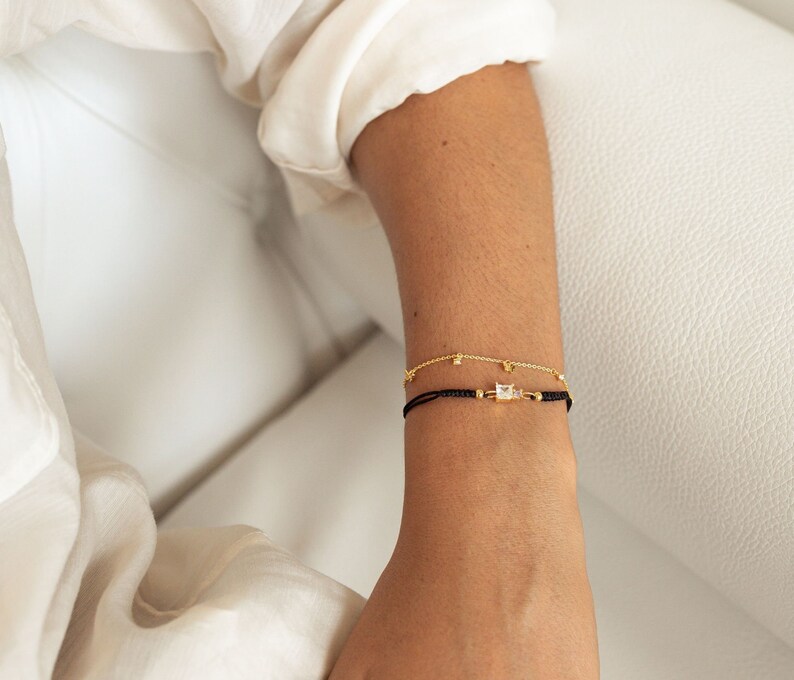 May include: Two bracelets on a wrist. The top bracelet is a delicate gold chain with small square-shaped stones. The bottom bracelet is a black string with a larger square-shaped stone in the center.
