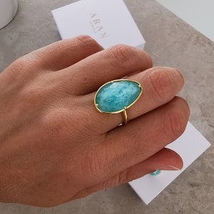 May include: A gold ring featuring a large oval turquoise gemstone. The ring is worn on a person's finger.