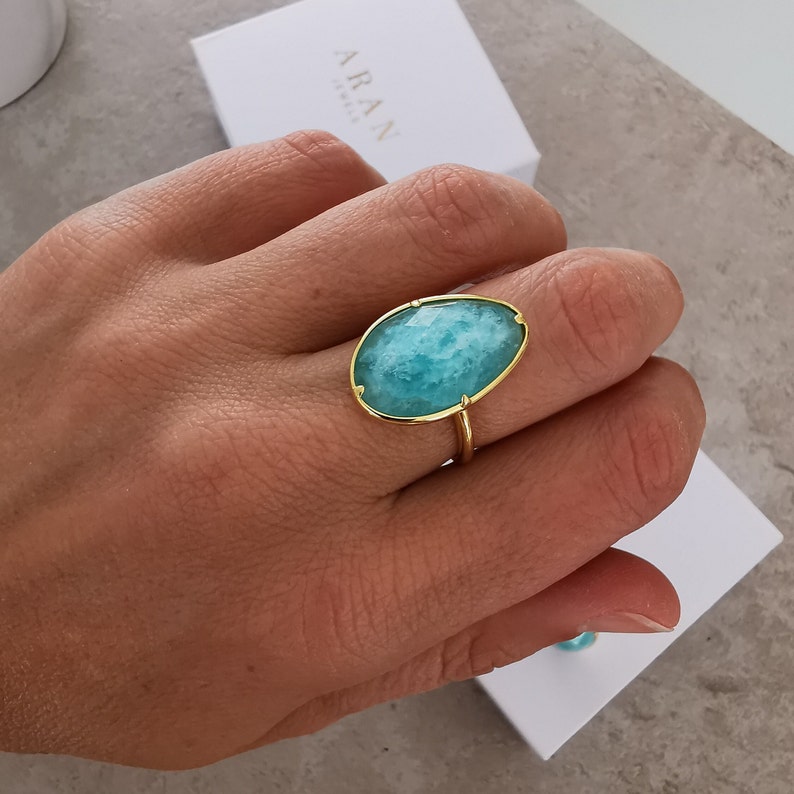 May include: A gold ring featuring a large oval turquoise gemstone. The ring is worn on a person's finger.