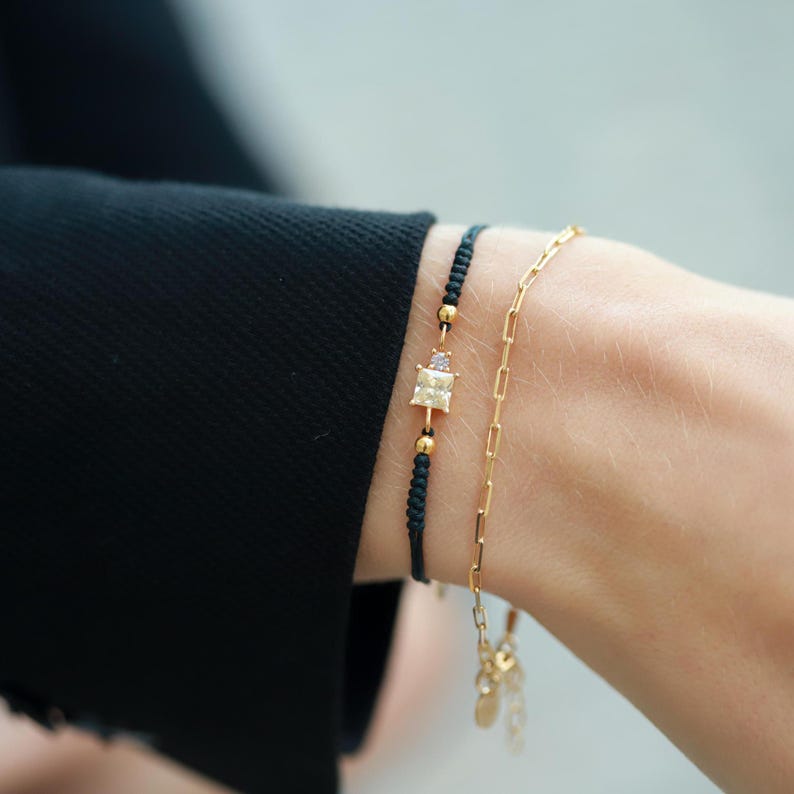 May include: Two bracelets on a wrist. One is a black string bracelet with a gold square charm with a clear stone. The other is a gold chain bracelet.