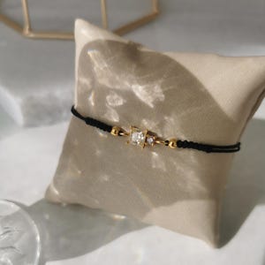 May include: A black string bracelet with gold accents and a clear square-shaped gemstone. The bracelet is on a light brown surface.