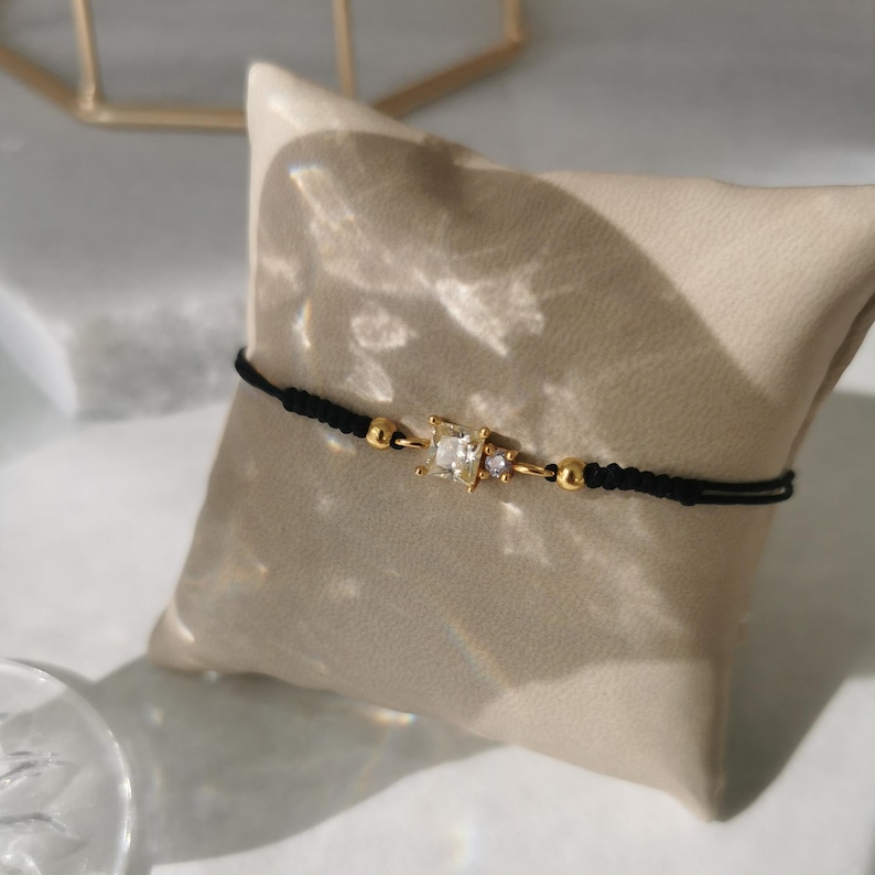 May include: A black string bracelet with gold accents and a clear square-shaped gemstone. The bracelet is on a light brown surface.