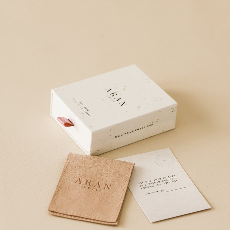 May include: A white box with a speckled pattern and the text "ARAN JEWELS" and "www.aranjewels.com". A brown suede pouch with the text "ARAN JEWELS" and a white card with the text "ALL YOU NEED IN LIFE IS A FRIEND WHO HAS CHOCOLATE... IT'S ME! with love for you"