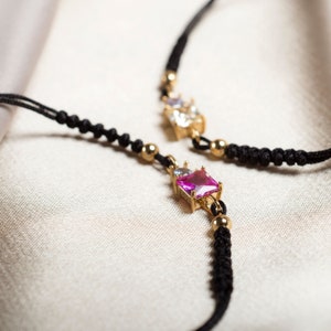 May include: A black cord necklace with a pink and clear gemstone pendant. The pendant is set in gold and has a square shape.
