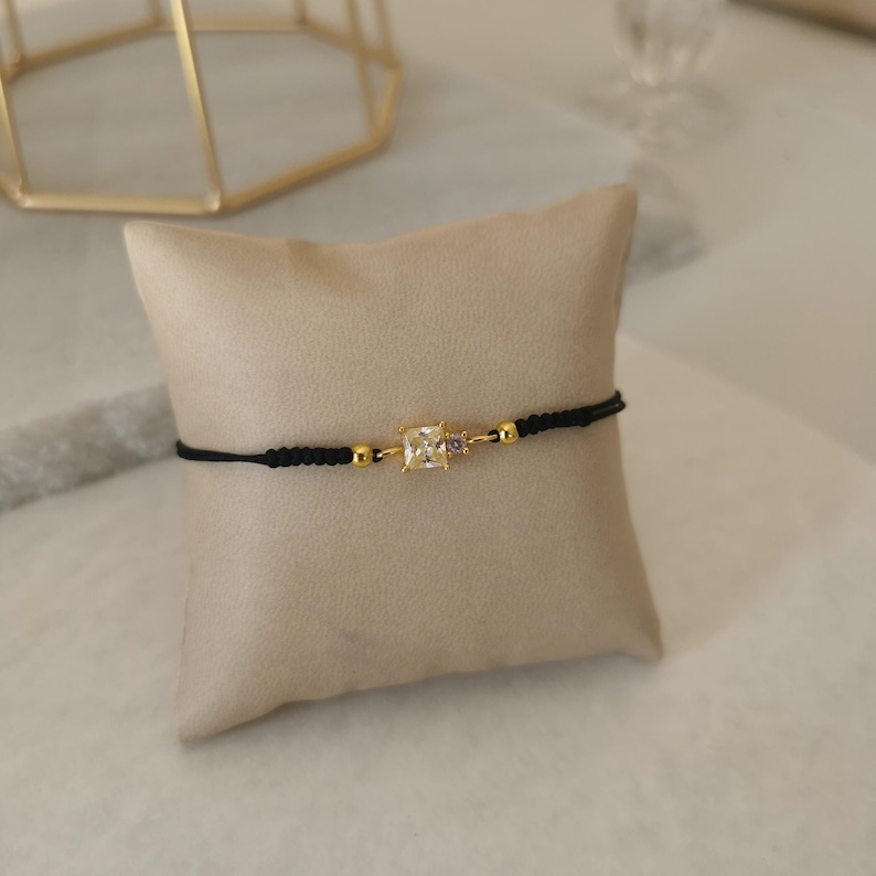 May include: A black string bracelet with a gold square-shaped charm featuring a clear crystal stone.