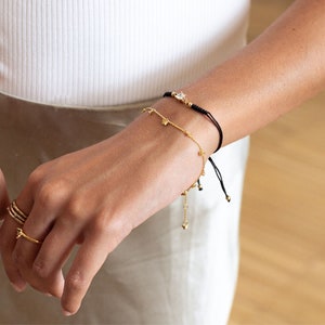 May include: A woman's wrist with three gold bracelets. The top bracelet is a black string bracelet with a gold charm. The middle bracelet is a delicate gold chain with small charms. The bottom bracelet is a gold chain with larger charms.
