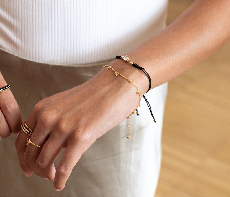 May include: A woman's wrist with three gold bracelets. The top bracelet is a black string bracelet with a gold charm. The middle bracelet is a delicate gold chain with small charms. The bottom bracelet is a gold chain with larger charms.
