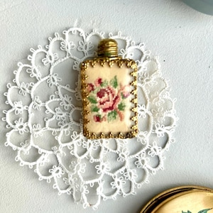 Puede incluir: Un pequeño frasco de perfume rectangular con tapón y ribete dorados. El frasco presenta un fondo color crema con un diseño floral bordado en rosa y verde. Descansa sobre un tapete de encaje blanco.