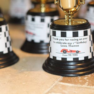 May include: A set of black and white checkered trophy cups with gold tops. Each cup has a personalized label with a red race car and the text "Thank you for racing on over to celebrate my 3rd birthday Love, Thomas June 20, 2015".