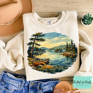 Könnte beinhalten: Cremefarbenes Sweatshirt mit einer malerischen See-Landschaft mit Boot, Bäumen und Bergen. Die Grafik ist in Blau-, Grün- und Gelbtönen gehalten. Das Sweatshirt wird mit blauen Jeansshorts, einem braunen Hut und Sandalen kombiniert.